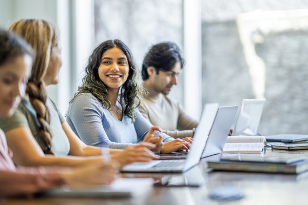 A group of four adult students sit together and study with one another. They have text books and laptops out in front of them as they take notes and talk amongst themselves.