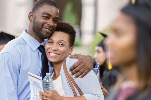 Two parents embrace as they watch their child in a graduation cap and gown from afar.