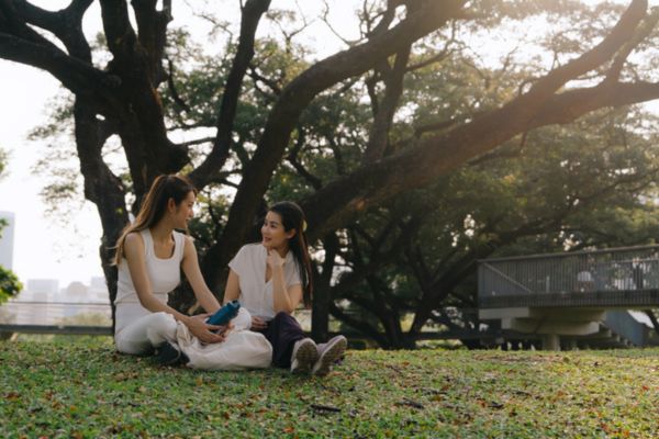Two women sitting and laughing together under a tree in a park.
