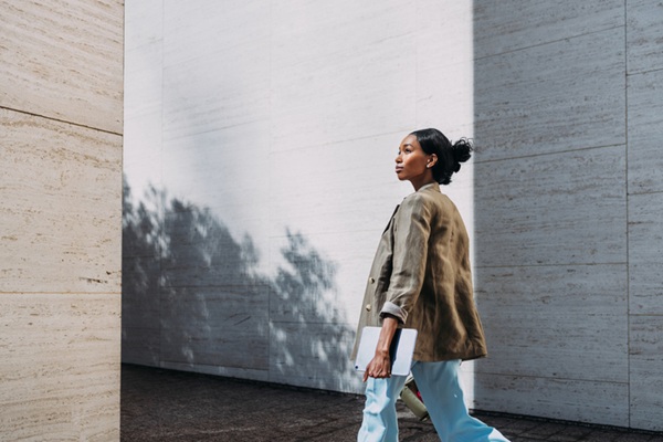A confident businesswoman in a casual blazer and trousers walks outdoors while holding a tablet, symbolizing professionalism and focus.
