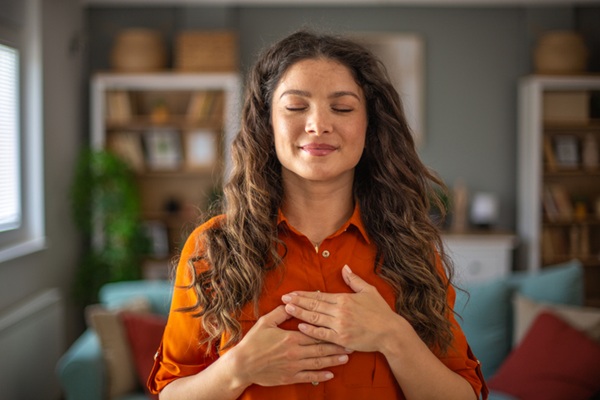 Young woman practicing mindfulness at home, holding hands on chest and enjoying peace.