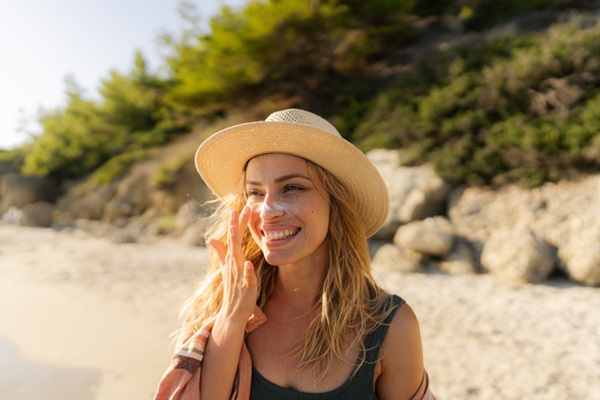 Young woman wearing a hat and applying sunscreen on her face at the beach.