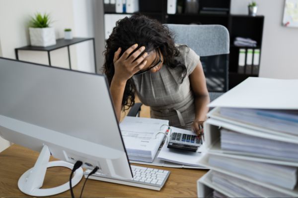 Stressed woman sits at a desk surrounded by files.
