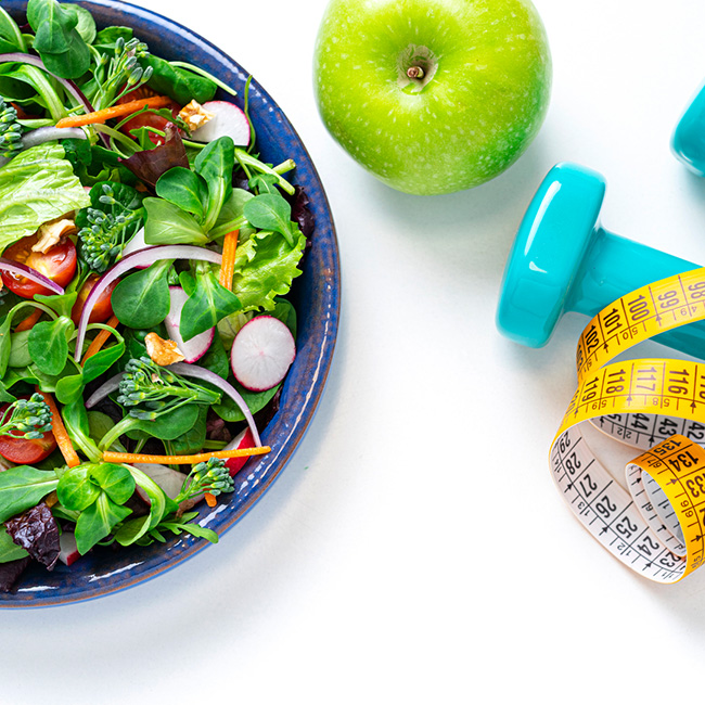 A bowl of salad, an apple, hand weights, and measuring tape